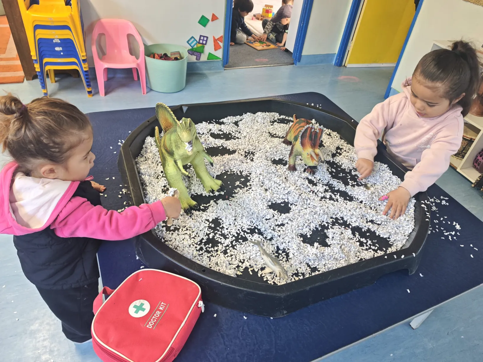 Two tamariki playing with dinosaurs in a sensory tray inside the preschool room
