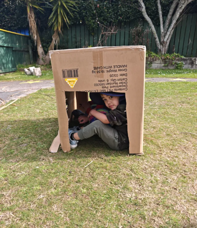 A tamaiti tucked inside a cardboard hut on the lawn