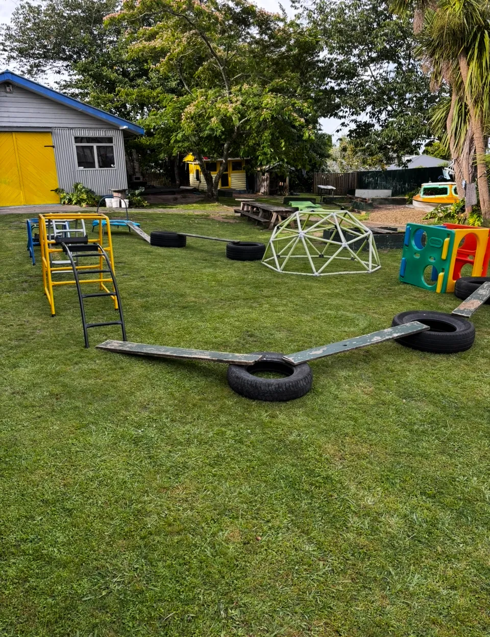 The big grassy backyard at Little Darlings Childcare in Rotorua, set up with tyres, planks, a climbing dome and play equipment