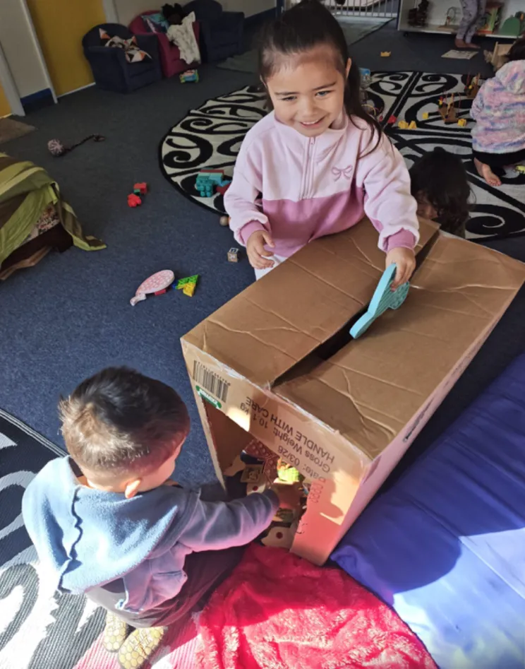 Two tamariki turning a big cardboard box into a house inside the play room