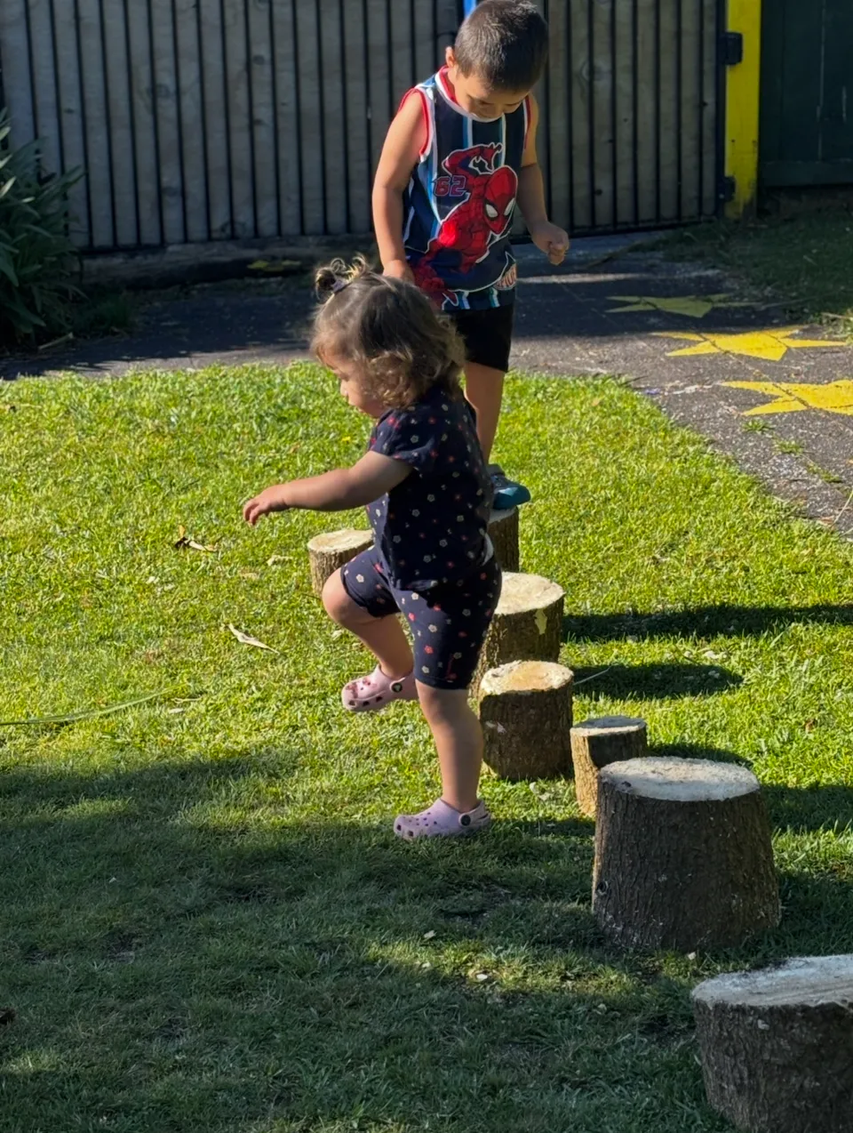 Two tamariki balancing across timber log steppers on the grass