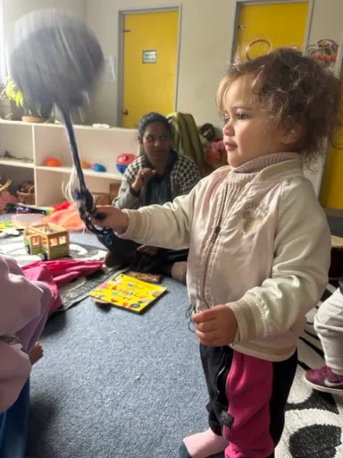 A kaiako sitting on the nursery floor while a toddler explores with a feather duster